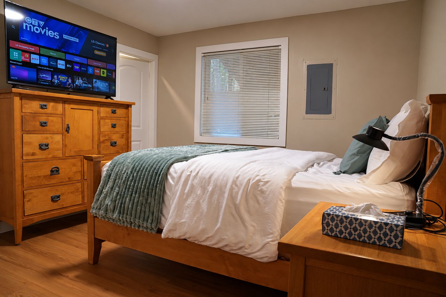 A bedroom with a bed, a wooden dresser, a television on the dresser, a window with blinds, and a bedside table with a lamp and tissues.