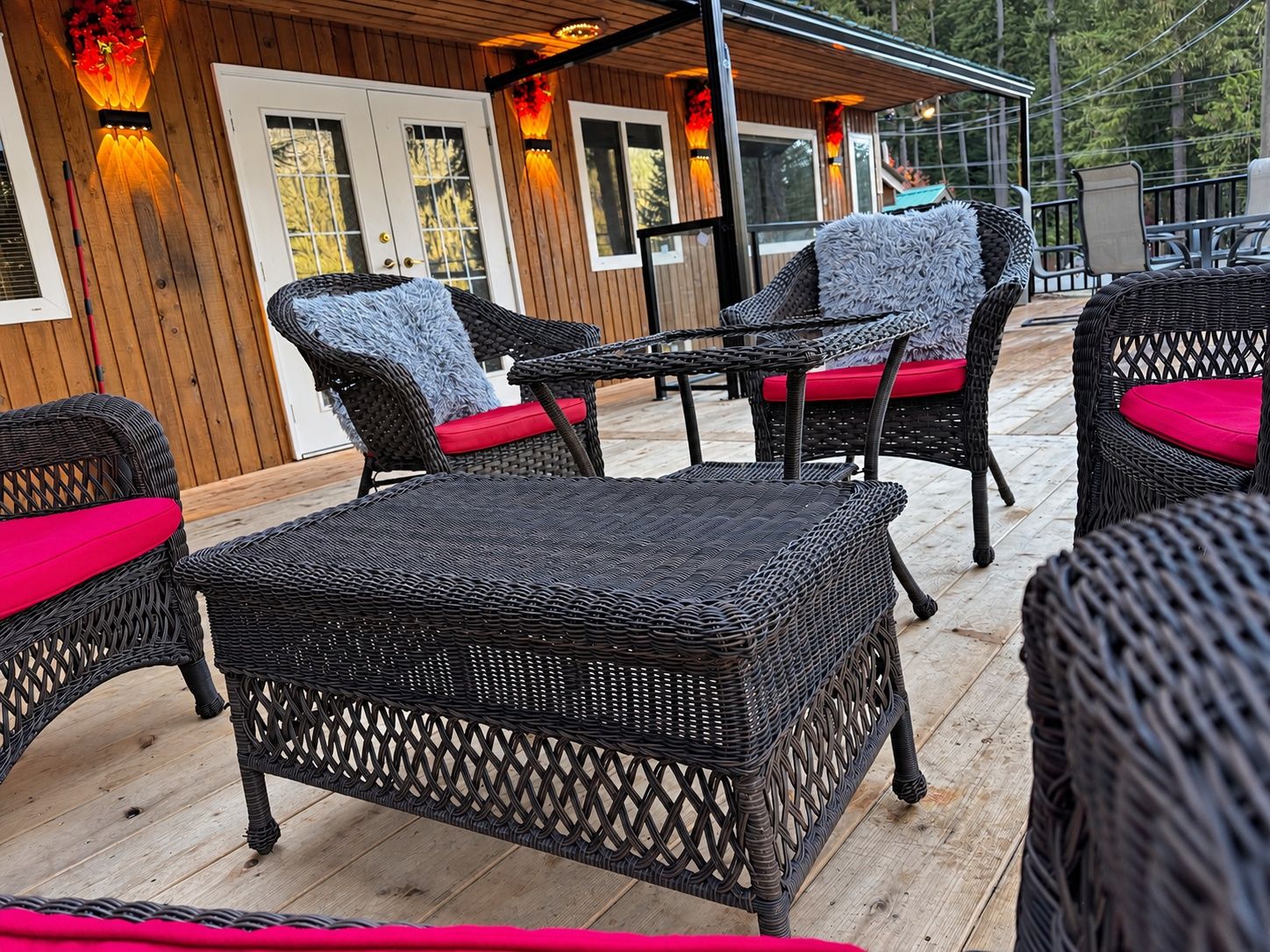 An outdoor patio area with wicker furniture and red cushions, wooden deck flooring, and a building with windows and doors in the background.