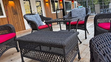 An outdoor patio area with wicker furniture and red cushions, wooden deck flooring, and a building with windows and doors in the background.