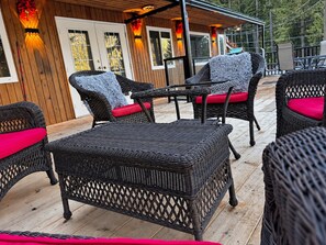 Een terras buiten met rieten meubels en rode kussens, houten dekplanken en een gebouw met ramen en deuren op de achtergrond.