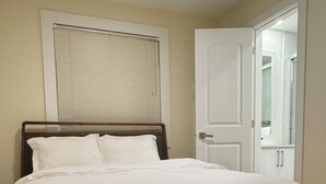 A bedroom with a wooden bed, white bedding, a window with blinds, and a white door.