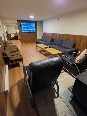 A hotel lobby with a wooden floor, a sofa, a black leather chair, a wooden coffee table, and a TV screen displaying a game.