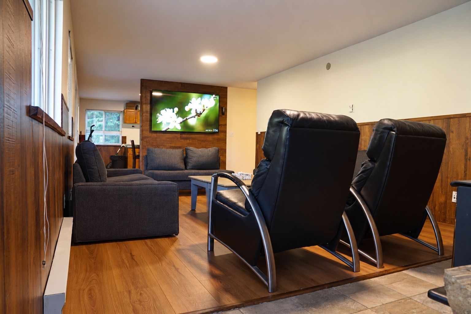 A modern living room with a sofa, two recliners, a coffee table, and a flat-screen TV mounted on a wooden wall.