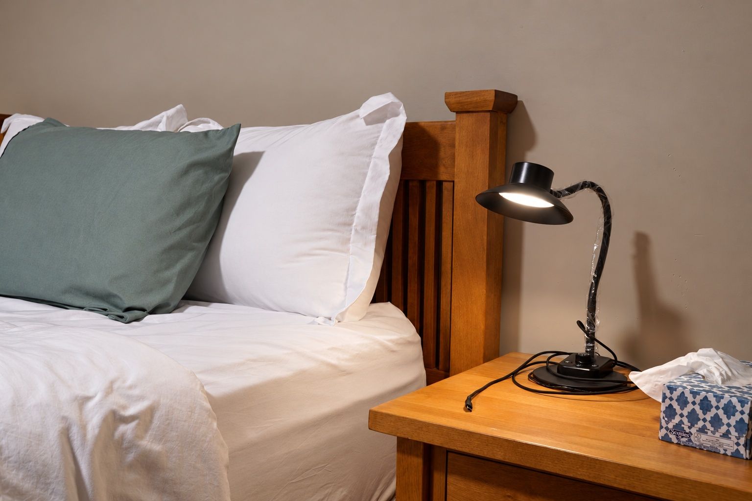 A neatly made bed with white and green pillows, a wooden headboard, a bedside table with a lamp, and a tissue box.