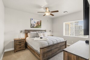 Une chambre à coucher comprenant un lit, des tables de chevet, un ventilateur de plafond, une fenêtre avec des stores et un tableau accroché au mur.