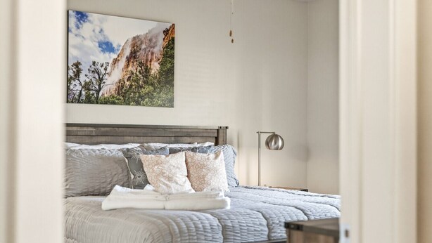 Ein Schlafzimmer mit einem Bett, Kissen, einer Decke, einem Nachttisch, einer Lampe und einem Bild an der Wand.