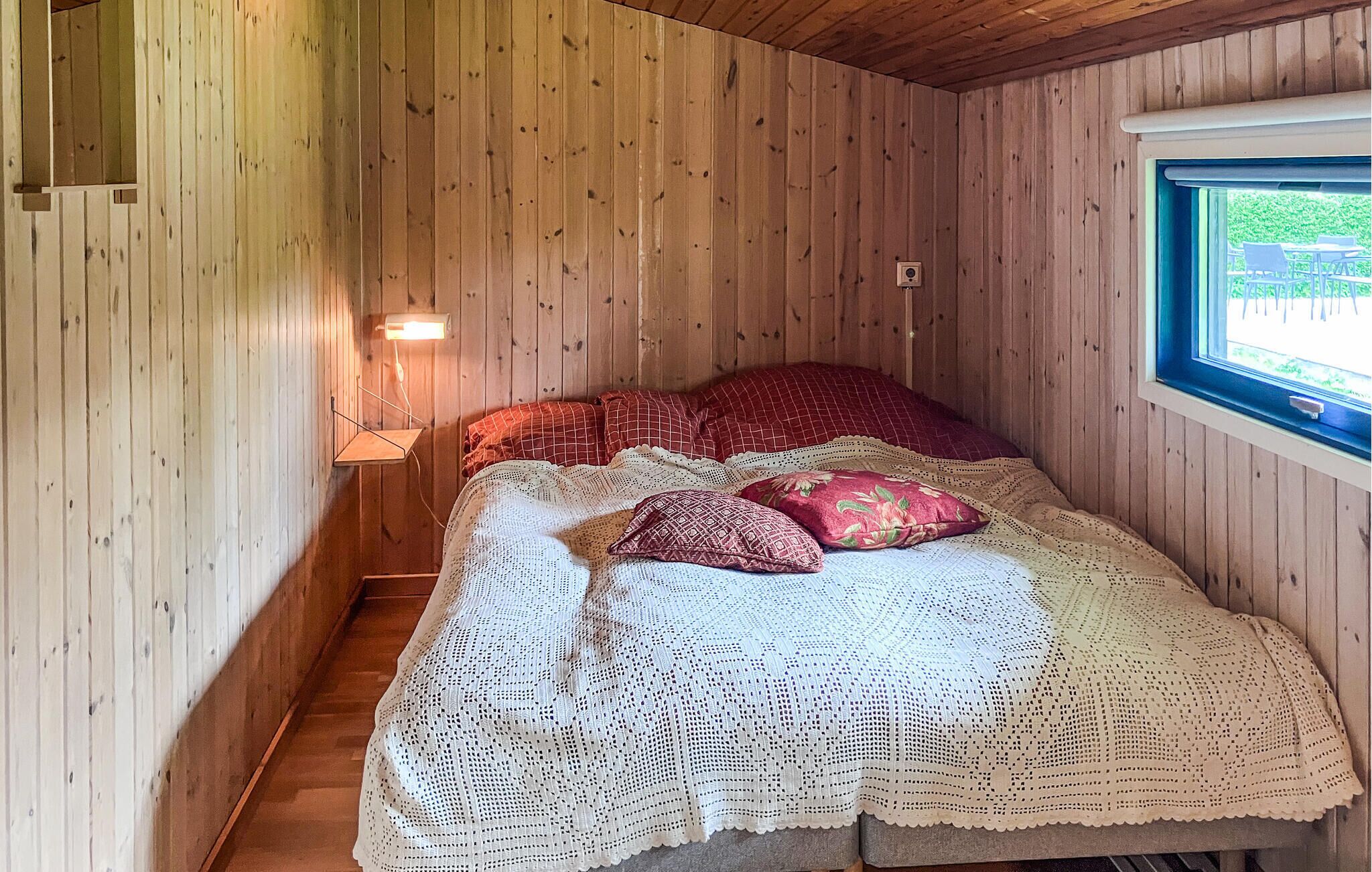 A cozy room with wooden walls, a bed with a white crocheted blanket, red pillows, and a window with a view.