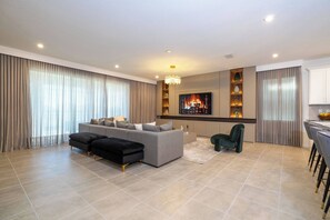 Ein modernes Wohnzimmer mit einem grauen Sofa, einem schwarzen Hocker und einem grünen Sessel. Ein Flachbildfernseher ist an der Wand montiert, darüber hängt eine Kristallleuchter, und es gibt eine Küchenzeile mit Barhocker.