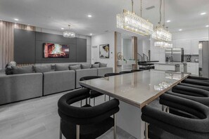 Eine moderne Küche mit Kochinsel, ein Essbereich mit Glastisch und ein Wohnbereich mit Sofa und Fernseher.