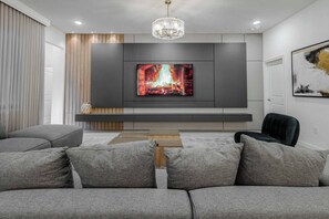 Ein modernes Wohnzimmer mit einem Flachbildfernseher an einer grauen Wand, einem Holztisch, grauen Sofas und einem schwarzen Sessel.