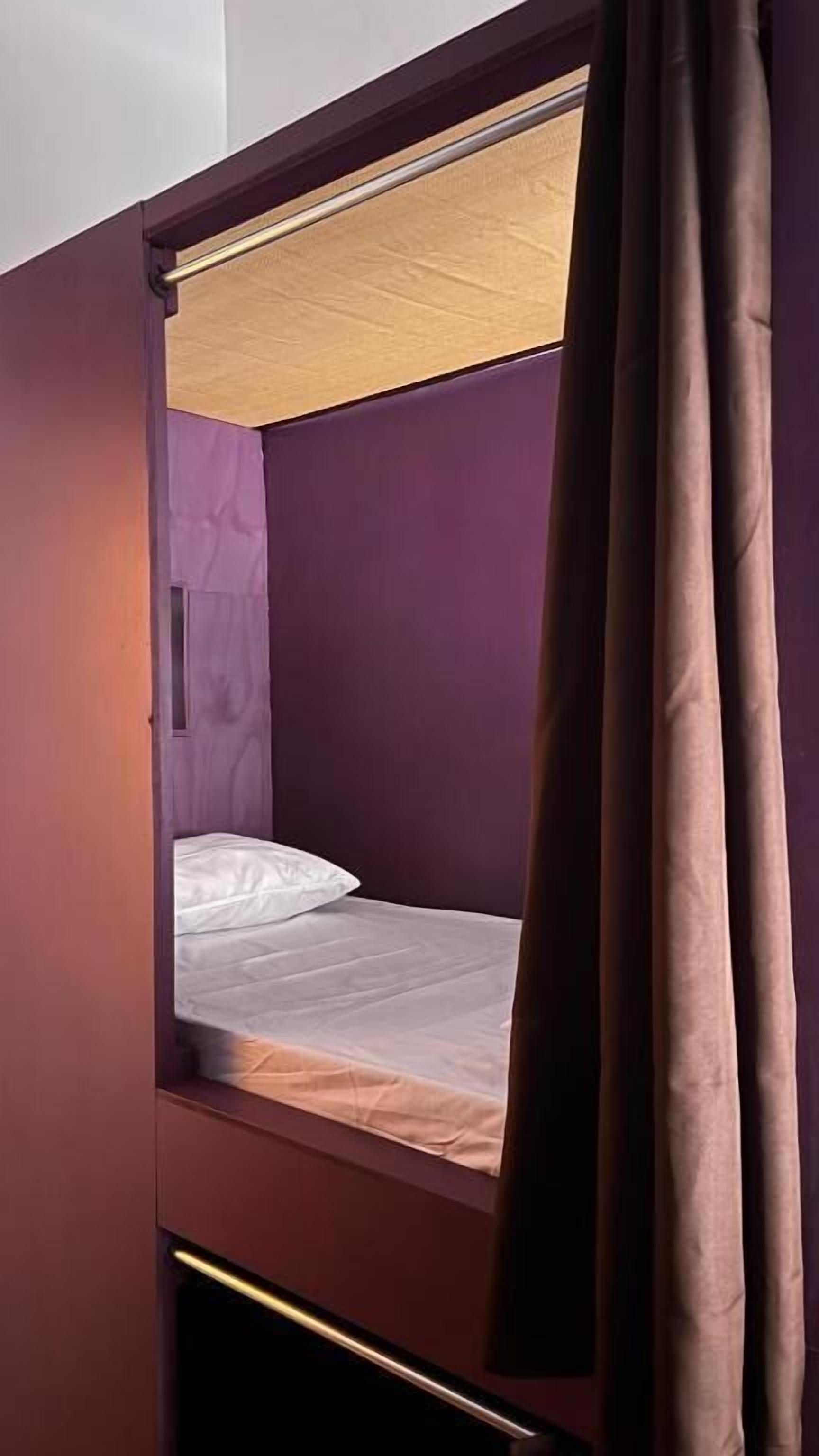 Una cama individual con dos literas, cabecera de madera, colchón blanco y cortina marrón.