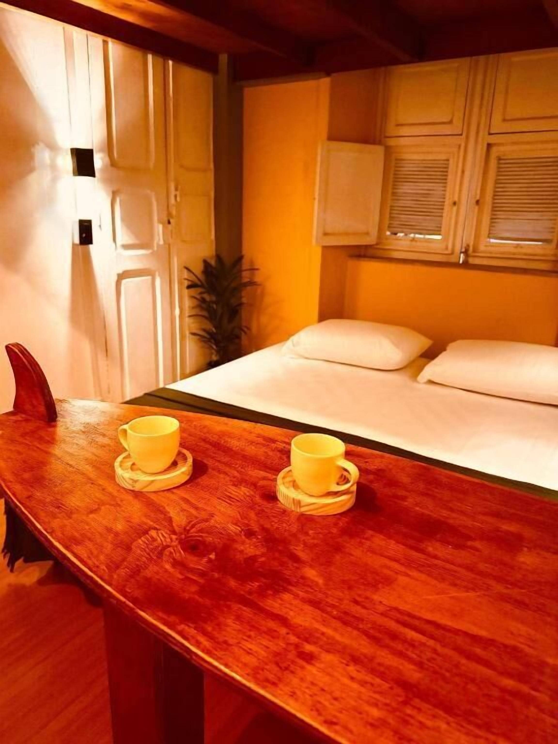 Una mesa de madera con dos tazas amarillas sobre posavasos, una cama con ropa blanca, una planta en maceta y una puerta de madera.