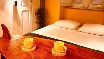 Una mesa de madera con dos tazas amarillas sobre posavasos, una cama con ropa blanca, una planta en maceta y una puerta de madera.