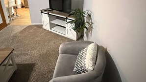 A living room with a grey sofa, a TV on a glass cabinet, a plant, and a kitchen in the background. - Cozy 2-bedroom in quaint Sturgis.  1 Mile to Main Street (Sturgis)