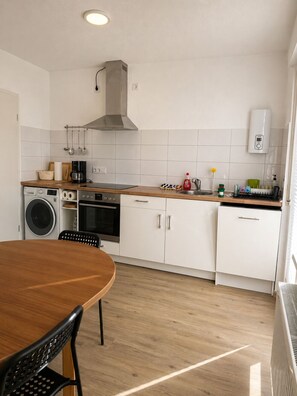 A compact kitchen with a wooden table, a washing machine, a coffee maker, and a built-in oven.
