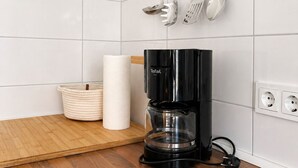Eine Küche mit weißen Fliesen, einer schwarzen Kaffeemaschine, einem Holztisch und hängenden Küchenutensilien.
