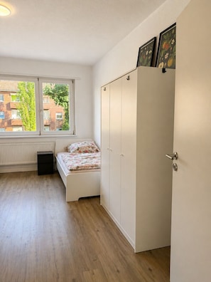 Ein Schlafzimmer mit einem Bett, einem Kleiderschrank, einem Heizkörper und einem Fenster mit Blick auf Gebäude.
