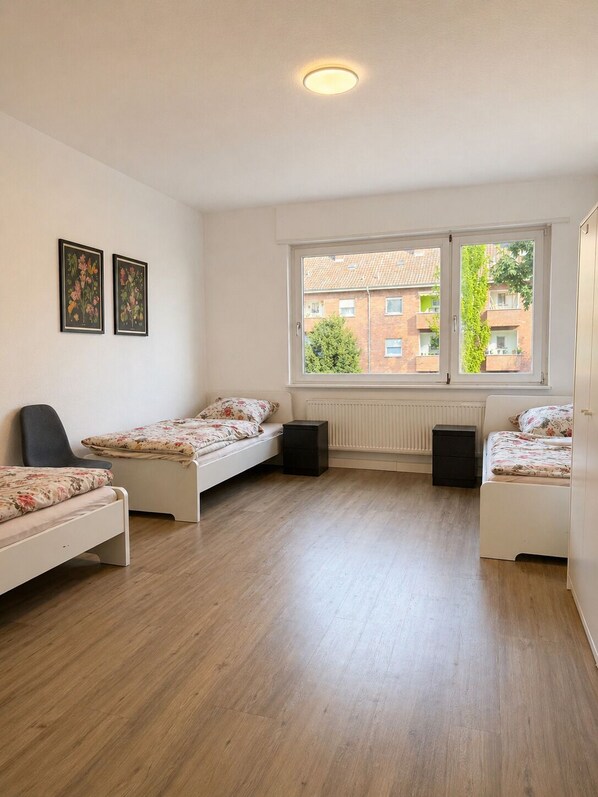 A bedroom with two single beds, a radiator, a window with a view of buildings, and two framed pictures on the wall.