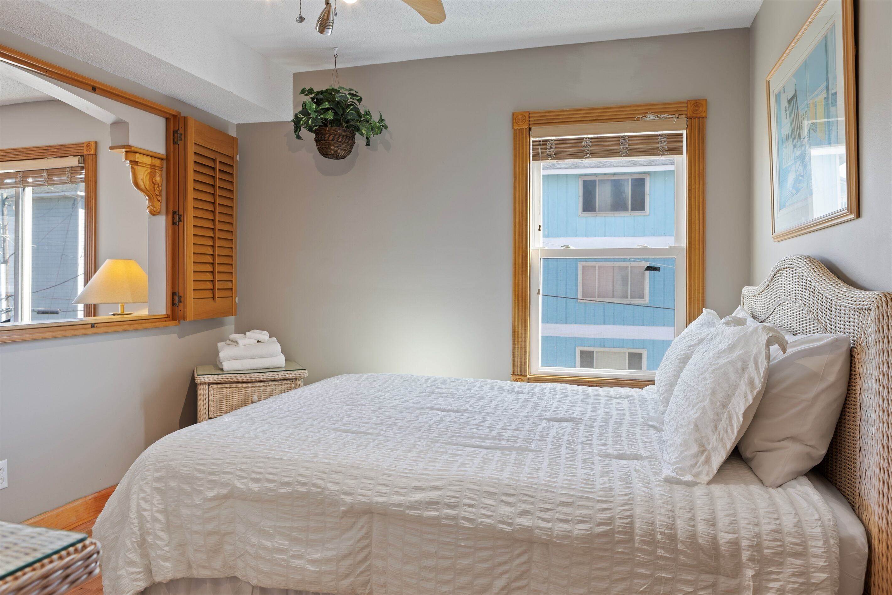 Ein Schlafzimmer mit Bett, Nachttisch, Lampe, Fenster mit Fensterläden und einer hängenden Pflanze.