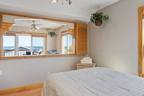 Ein Schlafzimmer mit einem großen Spiegel, Holzjalousien, einem Deckenventilator, einem Bett, einem kleinen Tisch mit Handtüchern und einer hängenden Pflanze.
