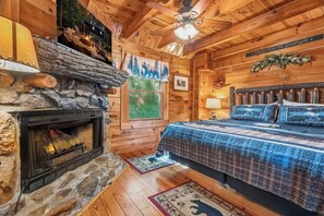 Ein Blockhaus-Zimmer mit Kamin aus Stein, einem Bett mit blau gemustertem Überwurf, einem Deckenventilator und einem Schild mit der Aufschrift 'Always Kiss Me Goodnight'.