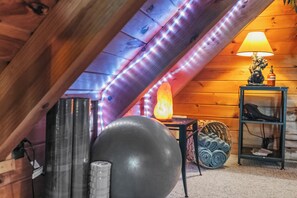 Ein gemütliches Dachzimmer mit Holzwänden, einer Lampe, Trainingsgeräten und einem kleinen Tisch.