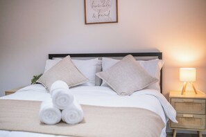 Ein ordentlich bezogenes Bett mit aufgerollten Handtüchern, einer Nachttischlampe und einem gerahmten Zitat an der Wand.