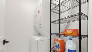 Un espace dédié à la lessive, avec un lave-linge blanc, une étagère en métal sur laquelle se trouve un détergent Tide, une bouteille d’eau de Javel et un rouleau d’essuie-tout.