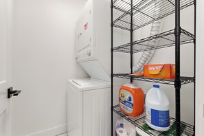 Ein Waschbereich mit einer weißen Waschmaschine, einem Metallregal mit Tide Waschmittel, einer Bleichmittel-Flasche und einer Rolle Küchenpapier.