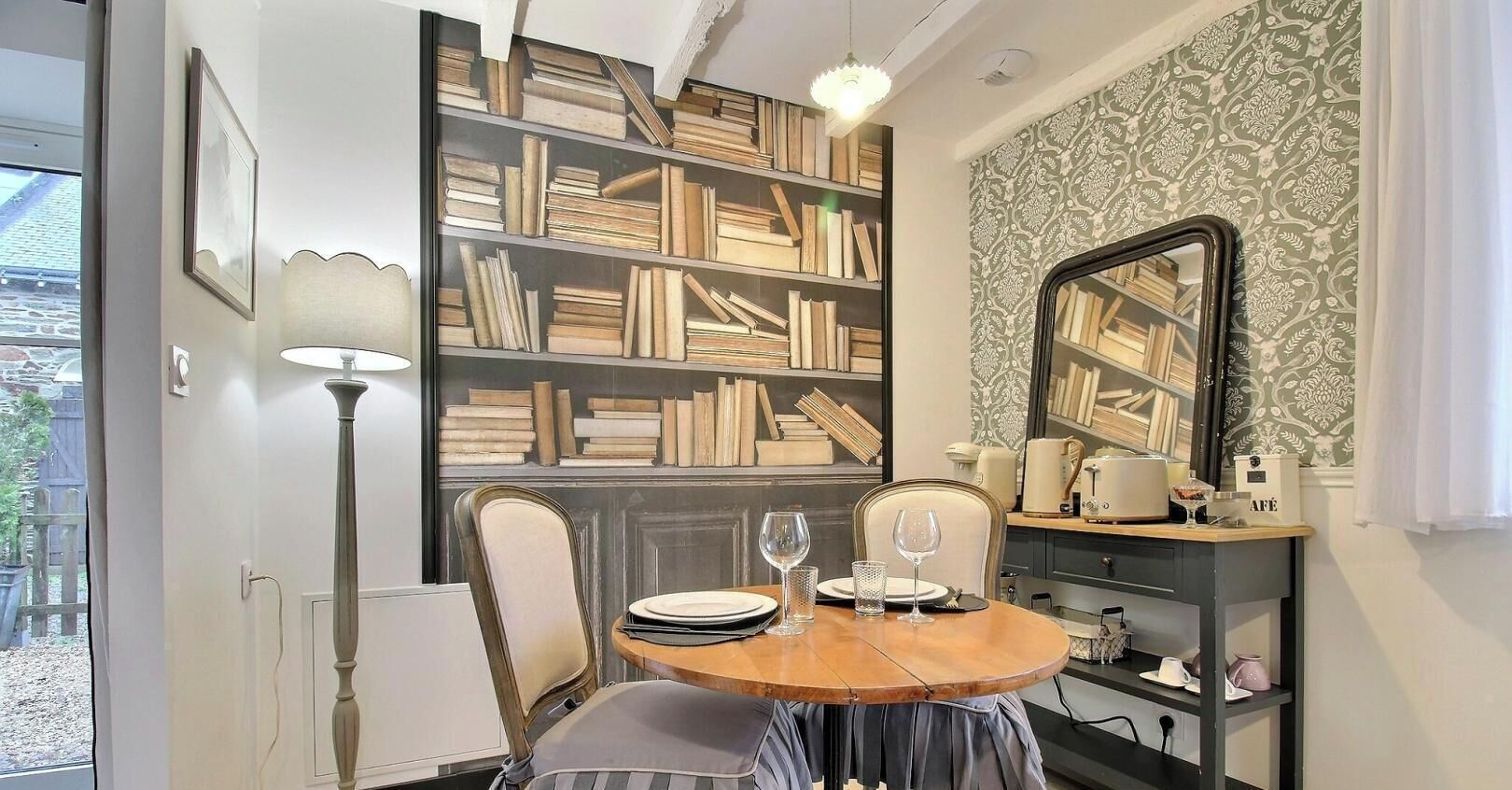 A dining area with a round table, chairs, a bookshelf, a mirror, and a window.