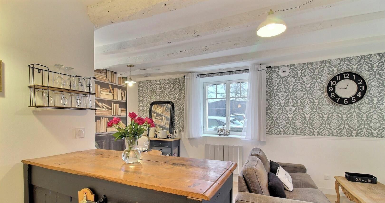 A room with a wooden table, a sofa, a clock, and a window with a view of the street.
