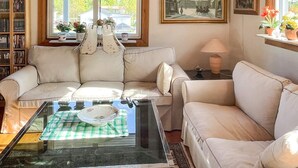 Un salón con una mesa de centro de vidrio, un sofá color beige, un tapete estampado, una estantería y un ventanal con vista a una casa.