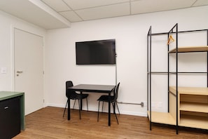 Ein Zimmer mit einem an der Wand montierten schwarzen Fernseher, einem schwarzen Tisch mit zwei Stühlen und einem Metallregal mit Holzablagen.