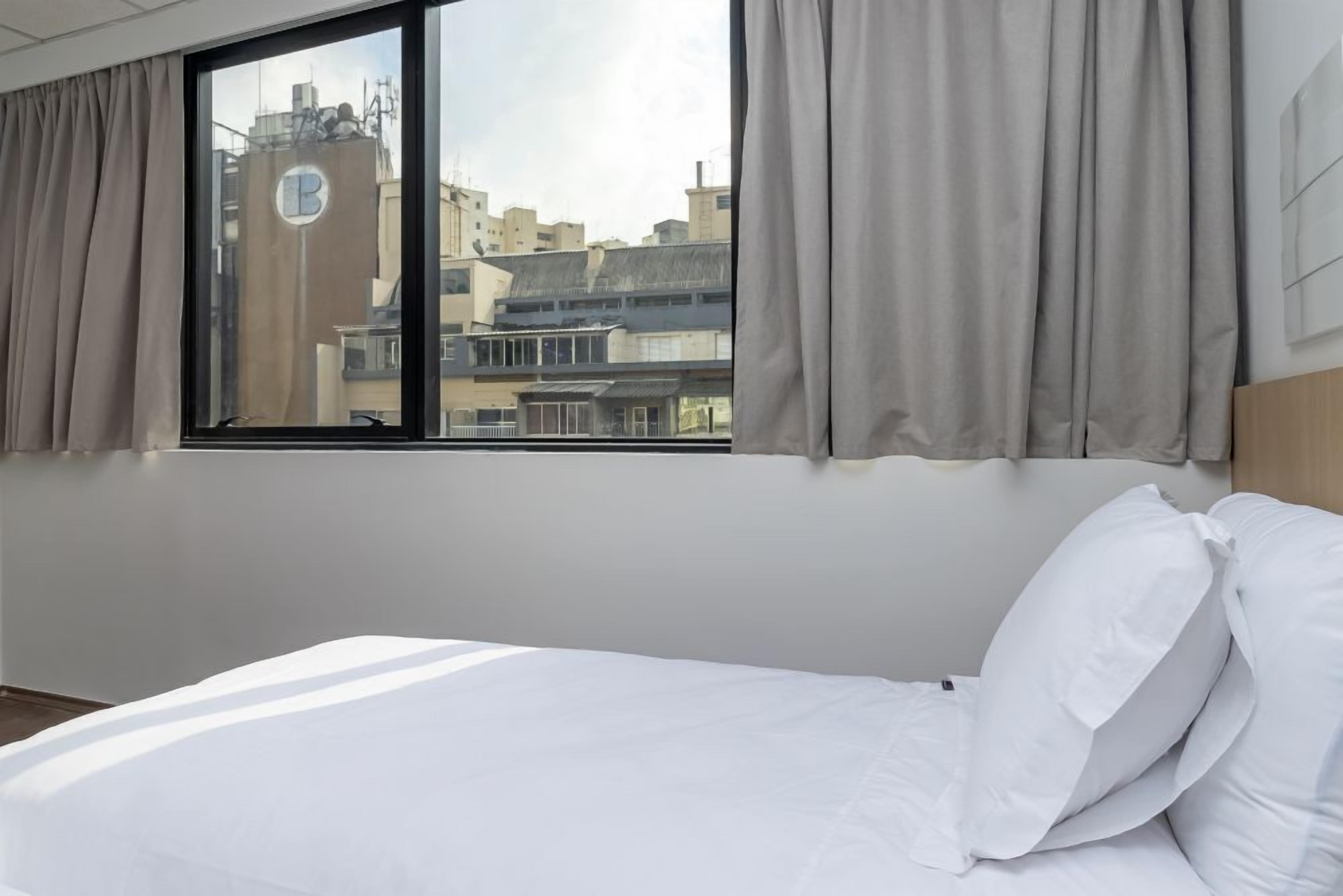 Habitación de hotel con cama, ventana con cortinas y vista a edificios.
