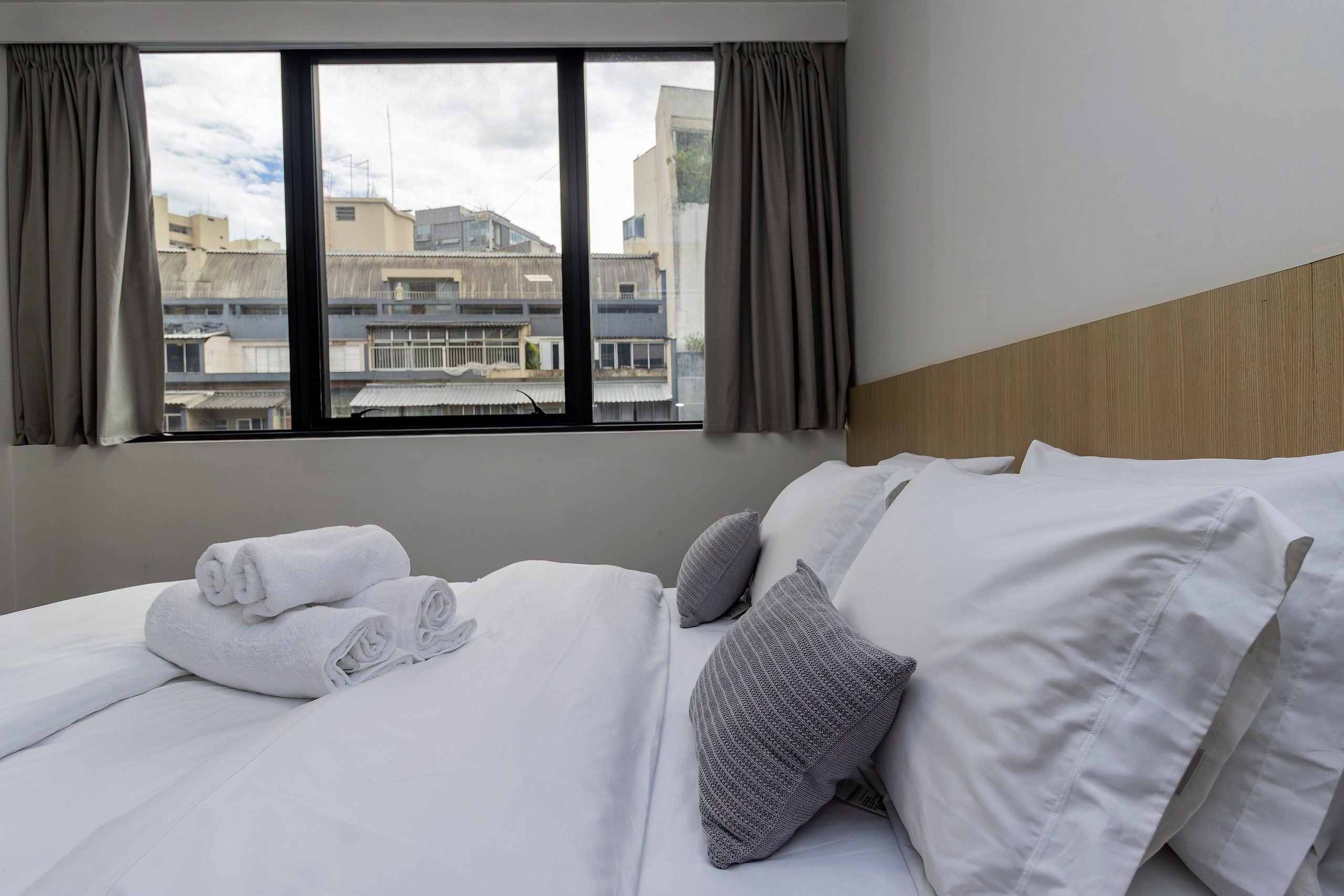 A hotel room with a bed, pillows, a window with curtains, and a view of buildings outside.