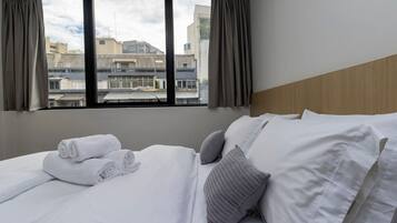 A hotel room with a bed, pillows, a window with curtains, and a view of buildings outside.