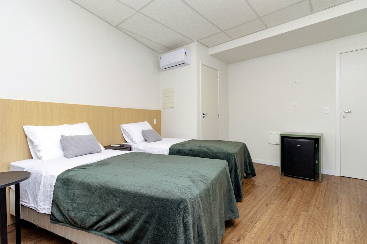 Ein Hotelzimmer mit zwei Betten, einem kleinen Tisch, einem Schreibtisch, einer Klimaanlage, einer Tür und einem Minikühlschrank.
