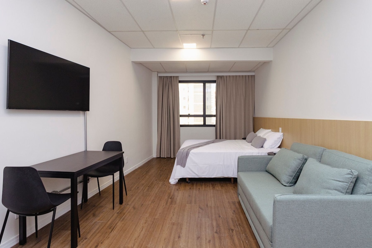 Ein Hotelzimmer mit Bett, Sofa, Schreibtisch mit Stuhl, Flachbildfernseher und einem Fenster mit Vorhängen.