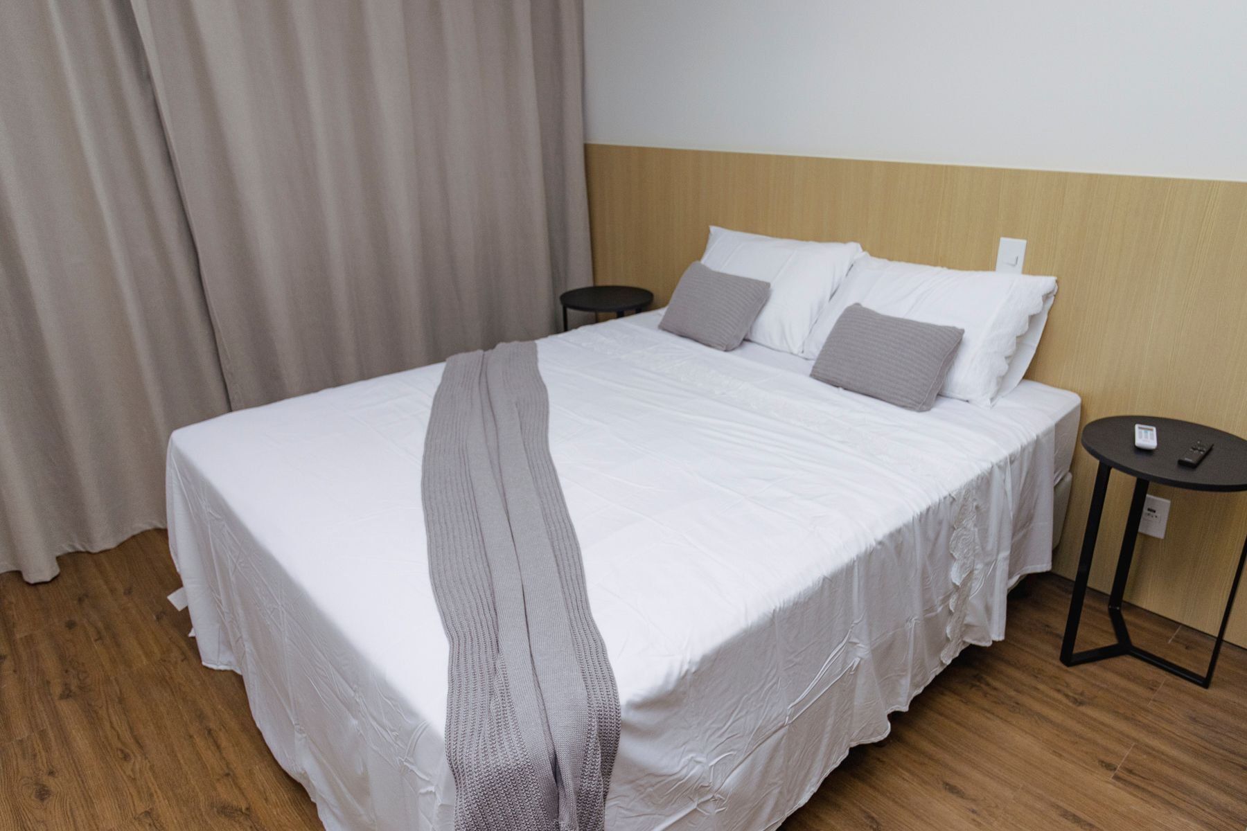 A neatly made bed with a grey blanket, two bedside tables with a remote control, and a curtain partially drawn.