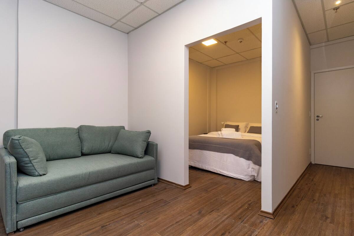 Um quarto com sofá verde, cama com roupa de cama branca e chão de madeira.
