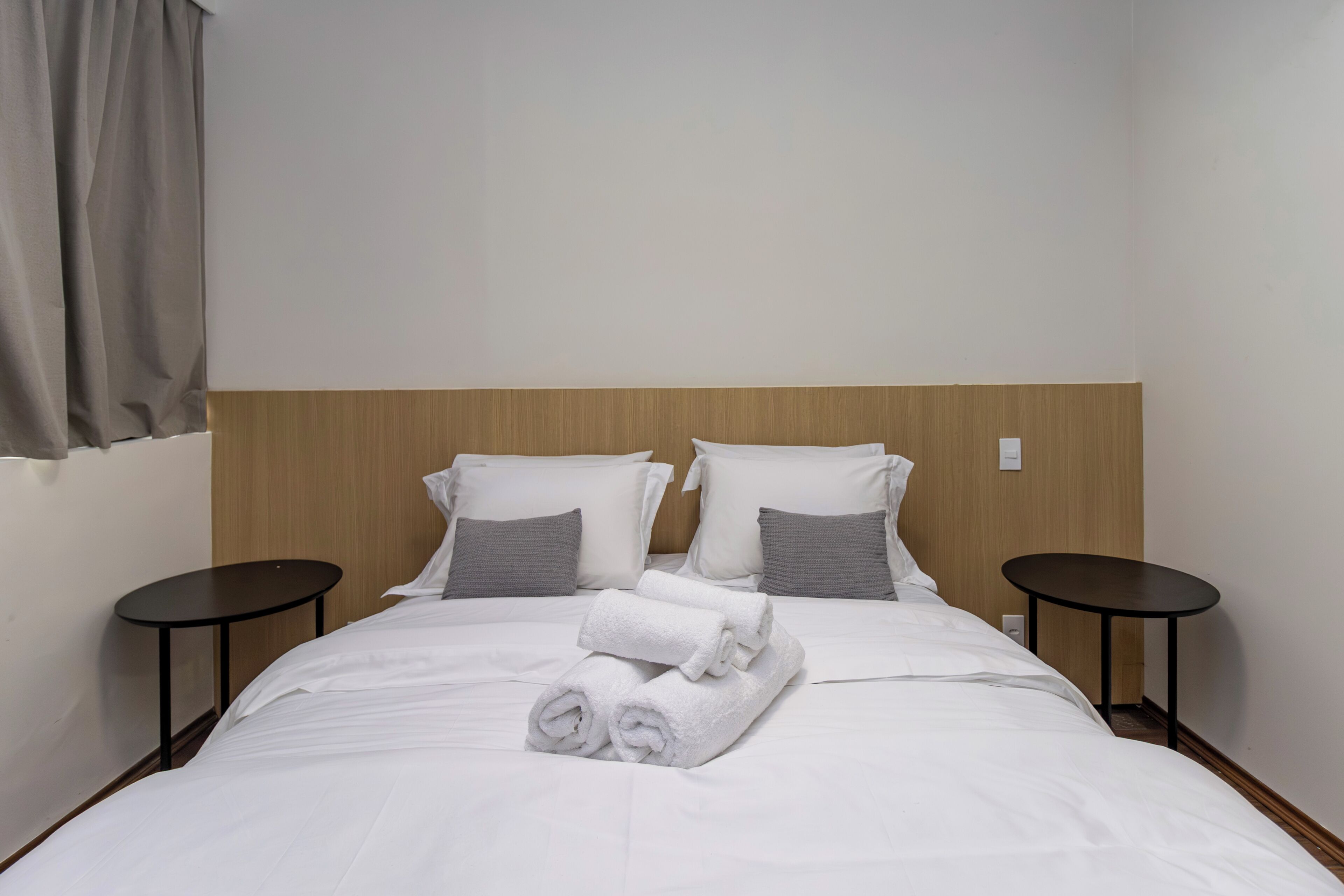 A neatly made bed with white linens and rolled towels, two bedside tables, and a wooden headboard.