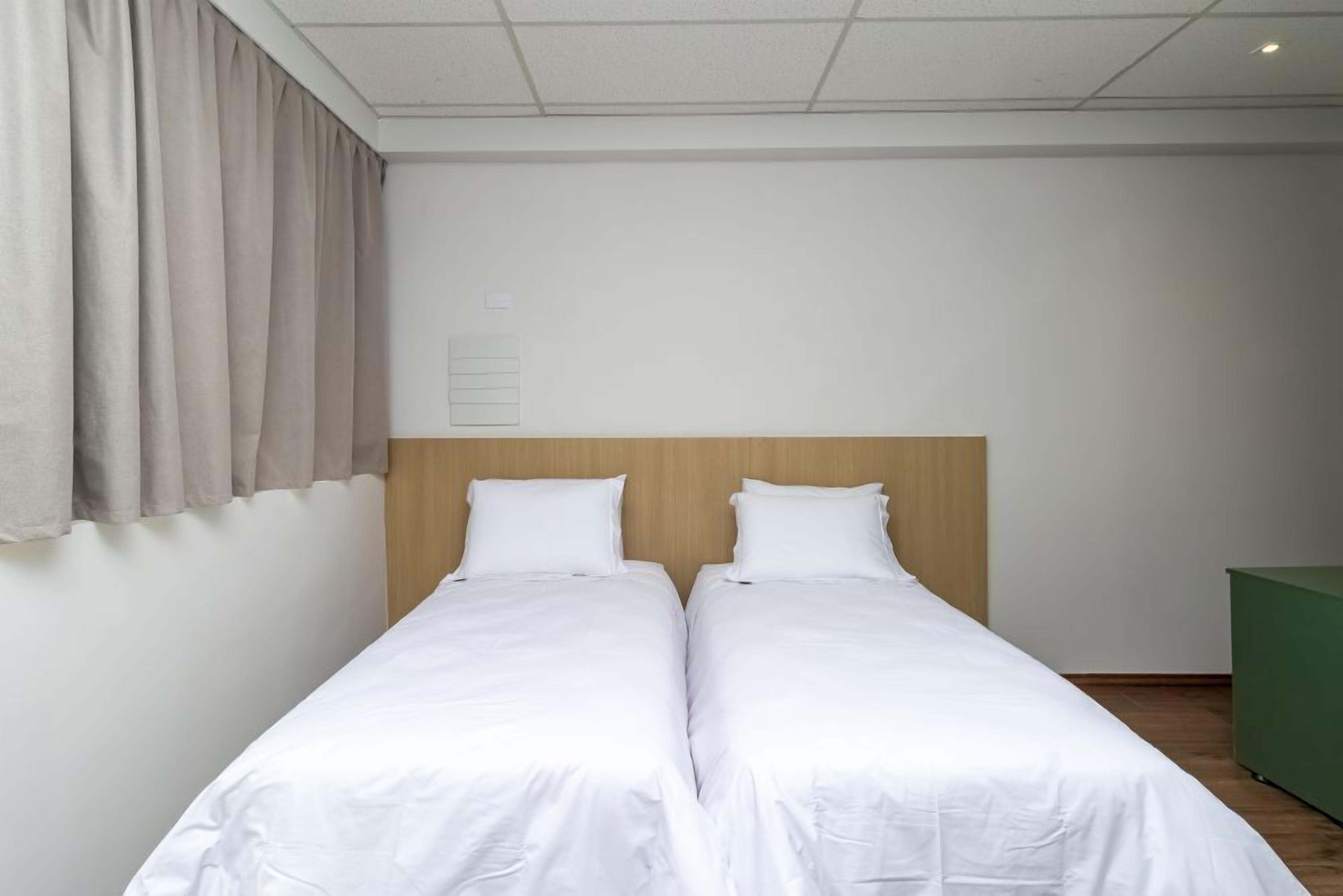 A double bed with white bedding, a wooden headboard, a green trash bin, and a window with curtains.