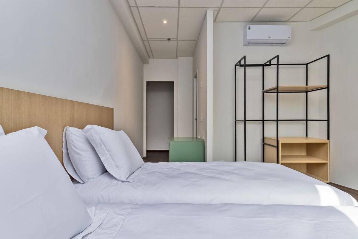 Quarto de hotel com cama, guarda-roupa, um cesto de lixo verde e uma estante de metal preta.