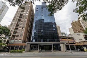 Um edifício moderno alto com fachada de vidro, rodeado por outros edifícios e árvores.