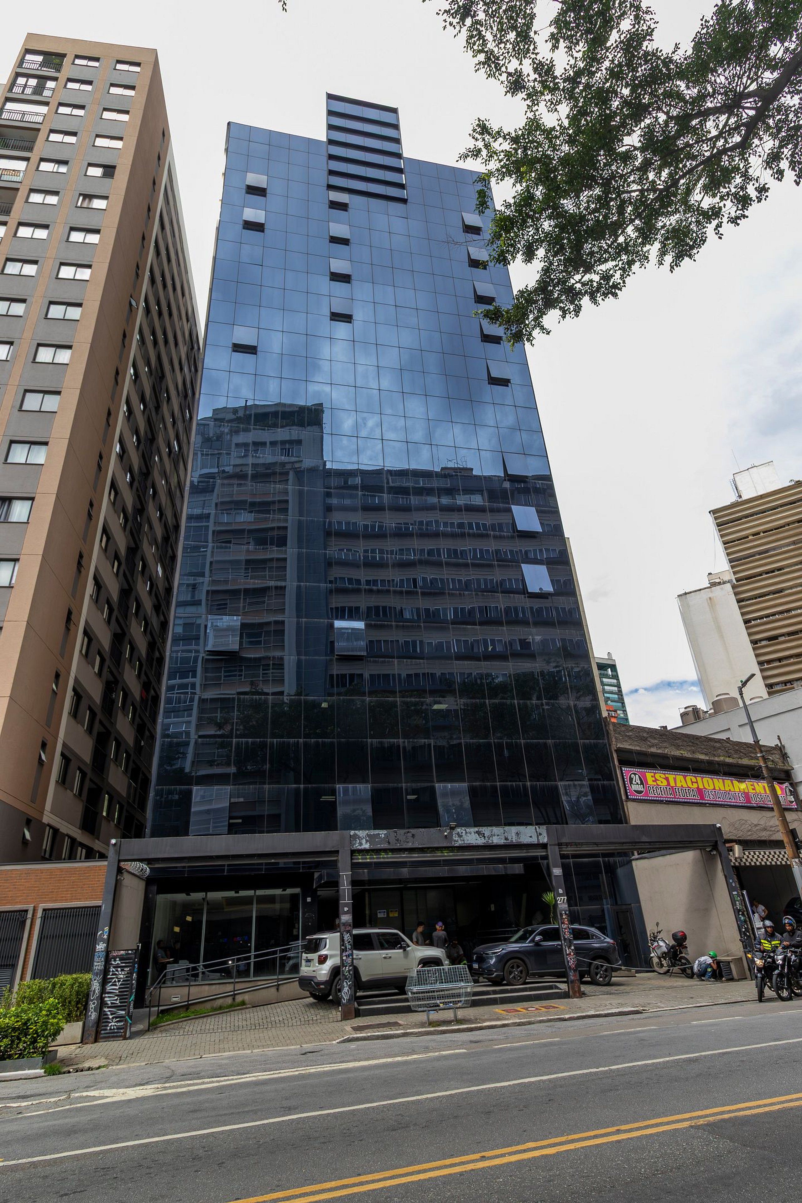 A modern high-rise building with a glass facade, a ground-level entrance, and a parking area with cars.