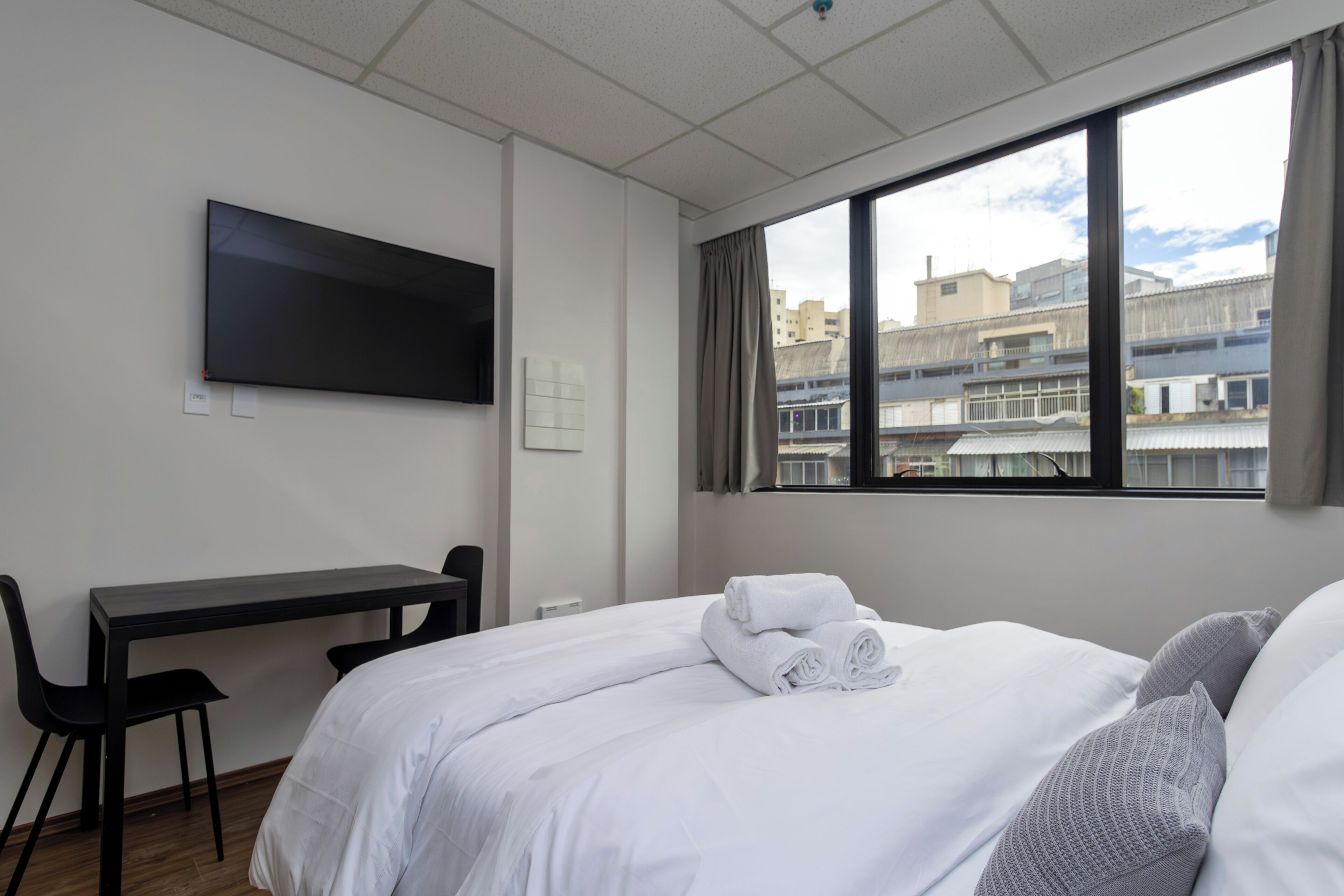 Ein Hotelzimmer mit Bett, Schreibtisch, Sessel, an der Wand befestigtem Fernseher und Fenster mit Blick auf die Stadt.
