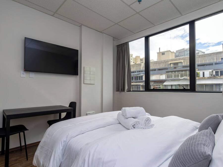 Ein Hotelzimmer mit Bett, Schreibtisch, Sessel, an der Wand befestigtem Fernseher und Fenster mit Blick auf die Stadt.