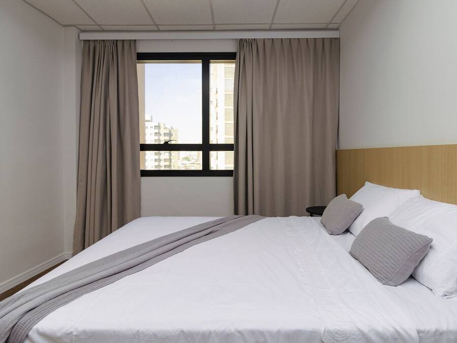 Una habitación de hotel con cama, cortinas, ventana y vista a edificios.