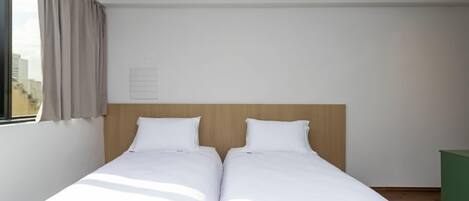 Una cama doble con cabecera de madera, un cubo de basura verde y una ventana con cortinas.