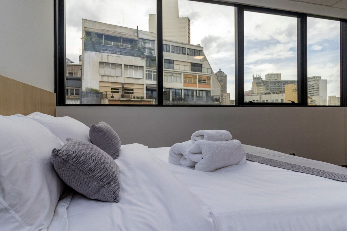 Ein Hotelzimmer mit Bett, Kissen und einem Fenster mit Blick auf die Stadt.